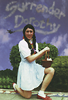 A man is kneeling on one knee dressed like blue gingham dress wearing a black pigtail wig holding a basket with both hands on his left knee. Written in grey writing on a blue sky are the words Surrender Dorothy. 