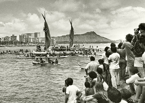 Black and white photograph, the Hōkūle‘a voyaging canoe coming into harbor after its inaugural voyage as spectators watch. 
