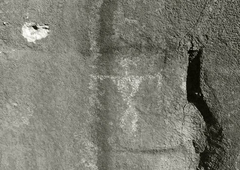 In this black and white photograph, a bullet hole is seen right above an ancient drawing of a person on a rock wall.