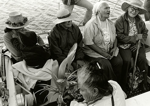Black and white photograph of a group of 4 elderly people and a younger woman holding a baby sit on a boat headed for Kaho‘olawe.