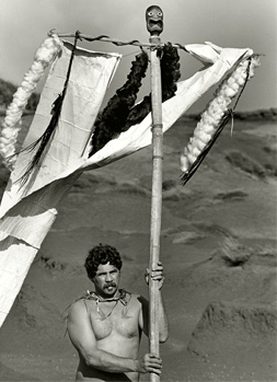 Black and white photograph of a shirtless man underneath a t-shaped pole made of wood with fabric draped across.