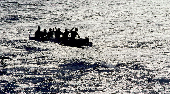 People on a canoe are in the middle of the sea. A small section of the sky can be seen above.