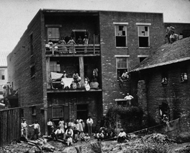 Black and white photograph of a brick building showing African American women on two balconies with hanging laundry. African American women and men along with white men and children can be seen sitting and standing in the courtyard below. Courtesy National Archives, Washington, D.C.