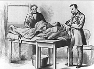 A young woman with diseased ovarian tissue (ovariotomy) lies on a table while a surgeon stands at her side with his left hand on her abdomen and his right hand near her knee. An anesthesiologist is preparing to administer an anesthesia.
