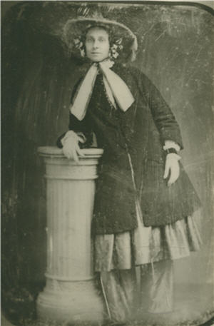 A woman leans on a pedastal to her right, wearing a brimmed hat and pants beneath a skirt and a coat.