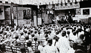 Black-and-white photo of a gathering attended by women and young girls. Raised stage in the front has a banner written in Chinese and its English translation, Health Pays Dividends. Underneath the banner, a person appears to address the gathering. The walls in the front of the room has multiple posters (indiscernible in the photo) on them.