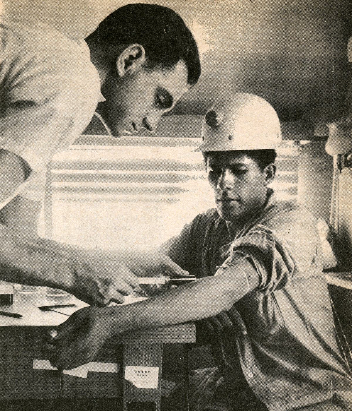 A white man standing leans down while taking blood from a seated Native American man in a hard hat.