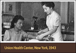 Standing White female nurse holds the arm of a seated African American woman.
