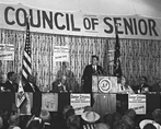 President Kennedy speaks at podium, flanked by 14 seated dignitaries, as a crowd looks on.