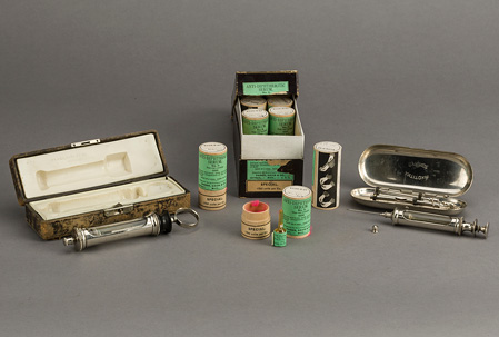 Box of wood canisters with green labels; small glass serum ampule is next to one open canister; two serum syringes with their cases are displayed on either side of box.