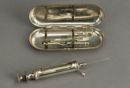 Metal serum syringe with needle attached is next to a metal case with hinged lid open.