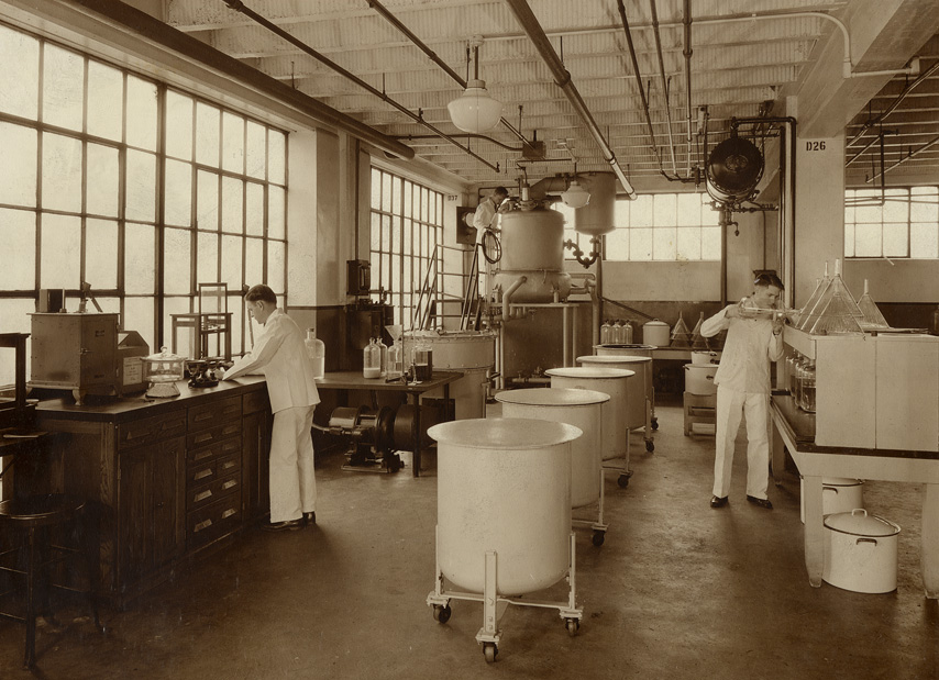 A man standing by a tall desk, and another man looking into one of four vats on wheeled frames.