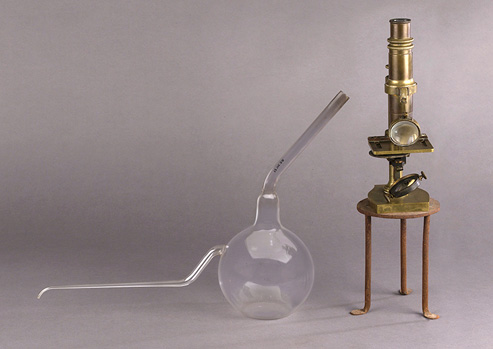 Bulb-shaped glass flask with two long thin necks sits next to an upright brass microscope on a small metal stand.