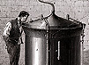A man stands next to a large cylindrical beer vat and gazes into the vat through a small opening in the conical cover. 
