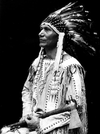 Dr. Charles A. Eastman seated left side pose, in traditional traditional Sioux war shirt with a feathered headdress holding a tomahawk.