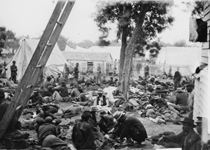 Black and white photograph a large group of men, some injured, sitting and lying on the ground in front of hospital tents.