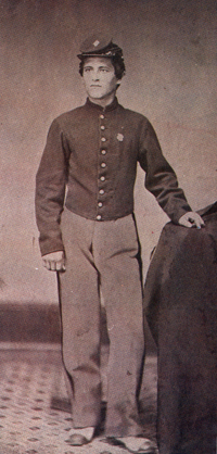 Sepia photograph of a uniformed Union soldier standing, left pose with his right hand resting on a covered table.
