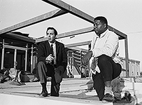 A white man and African American man kneeling observing a construction site.