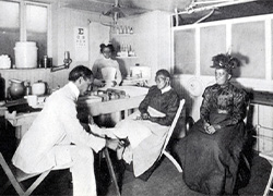 Physician seated in a chair tending to the leg of a seated patient with a nurse standing in the background.