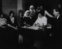 White woman in a headscarf completes a puzzle while seated. Two examiners and six women watch.