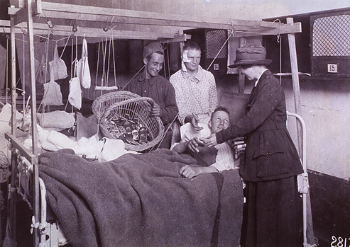 Photograph of two injured men in a hospital ward being handed cigarettes by a man and a woman.