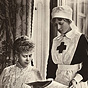 A White seated woman reads a newspaper with a White standing female nurse.
