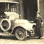 A White female nurse in dark military overcoat standing next to an automobile, looking at the viewer.