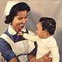 A Surinamese nurse in blue and white smiles at a Surinamese baby on her lap.