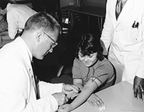 A photograph of a two white male doctors and a white female nurse drawing blood from two white girls