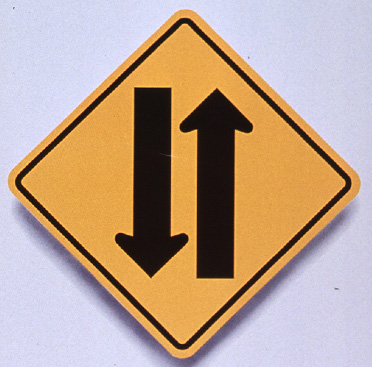 Yellow square road sign that has two vertical arrows pointing in opposite directions, above text