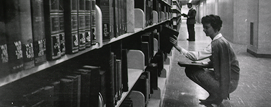 A woman kneels down to remove a book from the library stacks.