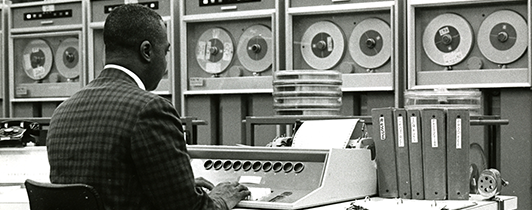 A man works at a keyboard surrounded by large tape data storage machines.