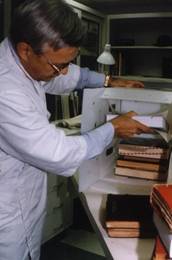 A man is packing a book into a plastic carton, which is sitting on its end.  He is stacking the books inside the bin with the spine edge facing the back of the bin. Freezer paper is between each book inside the bin.  Books waiting to be packed are sitting on the table by the bin.