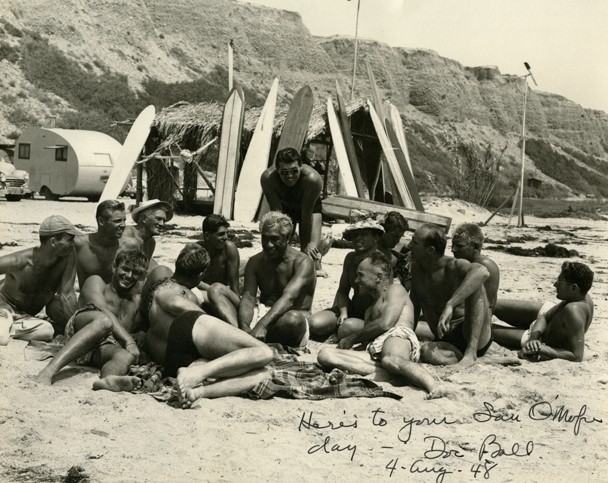 Black and white photograph of a group of surfers lounging in the sand as their surfboards lean against a thatched-roof structure in the background.