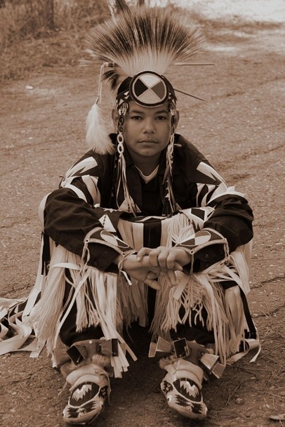 Sepia-toned image of a Native American boy wearing traditional regalia and a medicine wheel headband.