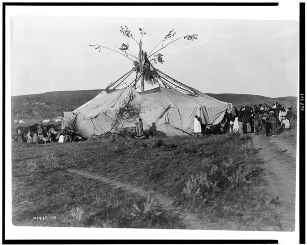 Black and white image of a group of Native individuals standing around a large, tent-like structure held up by tree branches.