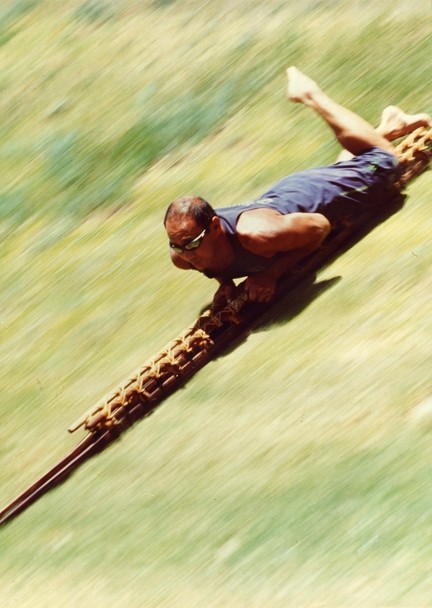 Color image of a male riding down a hill on a long, narrow, wooden sled.