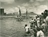 Black and white image of a group of onlookers standing on the beach, awaiting the arrival of a double-hulled voyaging canoe pictured in the background.