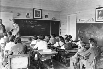 Black and white photograph of several Native American children at their desks inside of a one-room schoolhouse. 