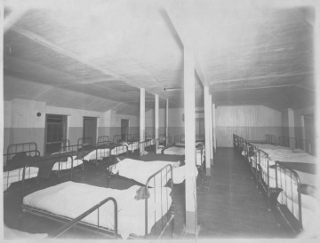 Black and white photograph depicting a large room with several rows of identical beds occupied by Native American boys.
