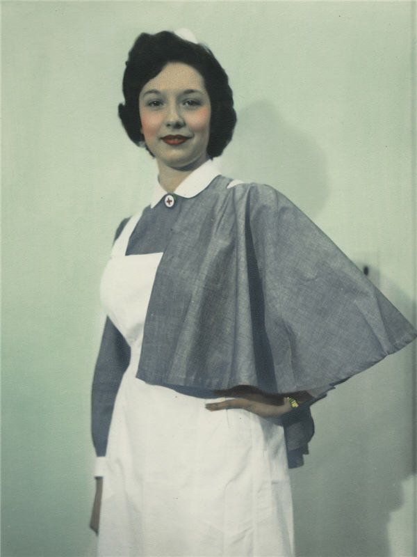 A posed photograph of a woman in a white cap and apron with an elbow length grey cape and a pin bearing a red cross.