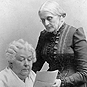 An old White woman sits and reads a pamphlet, another old White woman stands over her left shoulder.