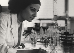 A woman leans toward a tall glass instrument on a laboratory desk.