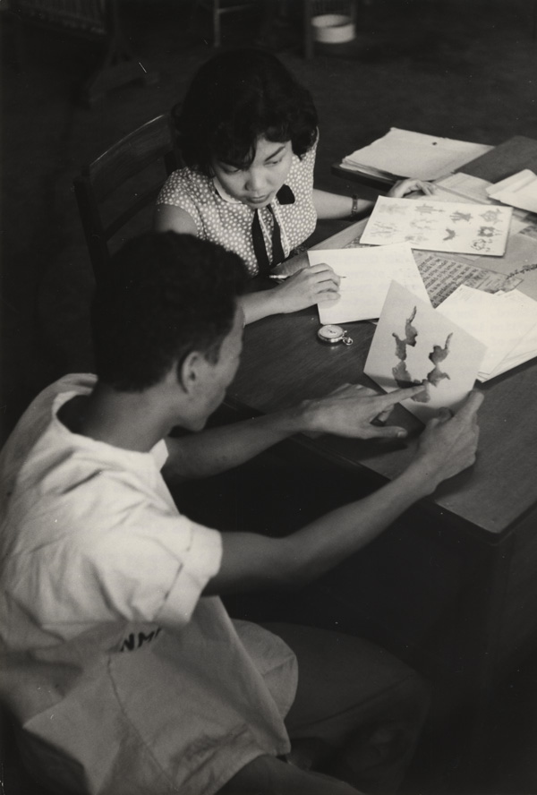 A woman observes a man seated to her right as he holds a card pointing to an image.  