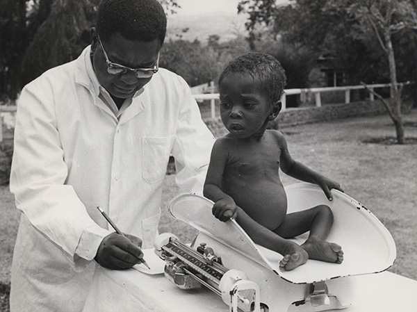 Outside, a physician weighs a child seated on a scale. 