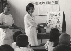 In front of a classroom, two women stand facing seated students as one points to a chart and the other stands on the side. 
