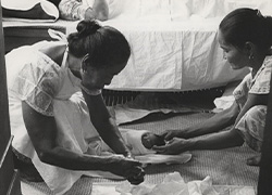 A woman lays in bed in the background; two woman sits across and over the matt they placed for an infant and supplies.