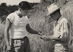 Outside, a man stands with his right palm open as another man holds a small bottle tilted over the palm. 