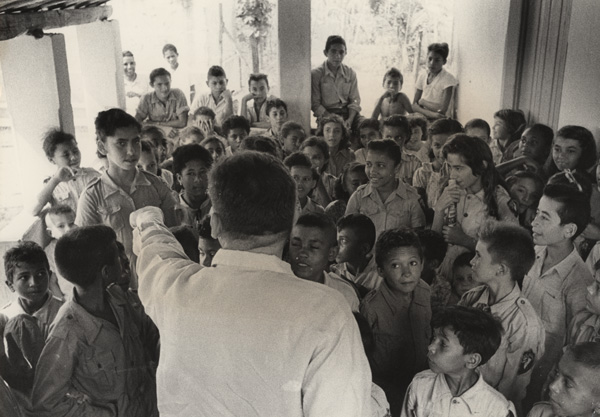 A large group of uniformed children are gathered around a health educator at a youth health club.