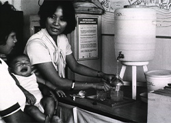 A woman shows water level in a bottle to fill as another woman looks on holding a crying baby.